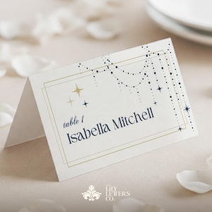 Puede incluir: Una tarjeta de mesa plegada con el nombre "Isabella Mitchell" en azul marino, con detalles de estrellas y puntos dorados. La tarjeta es de color crema con un borde dorado y el texto "mesa 1". El logotipo de The Lily Letters Co. está en la parte inferior.