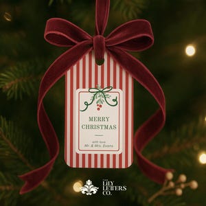 Può includere: Un'etichetta regalo natalizia a righe rosse e bianche con un nastro di velluto bordeaux. L'etichetta ha un bordo bianco con agrifoglio e le parole "MERRY CHRISTMAS". È appesa a un albero di Natale.
