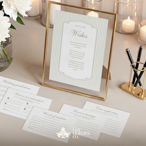 May include: A wedding decor setup with a gold-framed sign reading "Advice and Wishes" and lined paper. Several cards with prompts like "Wedding predictions" and "Words of wisdom" are scattered around. Black pens in a gold holder and white flowers complete the scene.