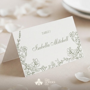 May include: White wedding place card with a floral design and the text "TABLE 1" and "Isabella Mitchell".