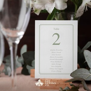 May include: A wedding table number card with a green border and text. The number "2" is displayed, with a list of names below. The card is set on a wooden base, with the words "LILY LETTERS CO." at the bottom.