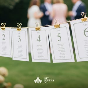 May include: White table cards numbered 1 to 6, suspended by gold clips on a string. Each card features a decorative border and guest names. The background is a blurred green, suggesting an outdoor wedding or event.