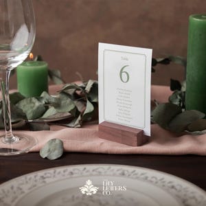 May include: A wedding table setting featuring a place card with table number 6, a green candle, and a wine glass. The place card is in a wooden holder. Eucalyptus leaves and a plate are also visible.
