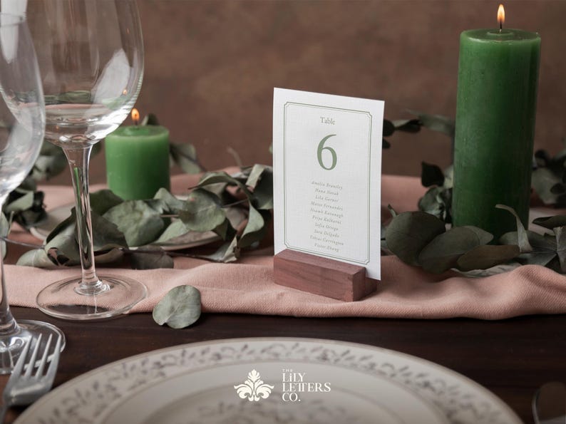 May include: A wedding table setting featuring a place card with table number 6, a green candle, and a wine glass. The place card is in a wooden holder. Eucalyptus leaves and a plate are also visible.