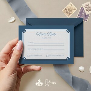 May include: A blue RSVP card with a white border and a decorative frame. The text on the card reads "Kindly Reply by April 17th". There are lines for the guest's name, dietary requirements, and two checkboxes for accepting or declining the invitation.