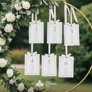 May include: A circular gold metal frame decorated with white flowers and foliage, with six white cards suspended from it. Each card is numbered and lists names. The cards are tied with white ribbons. The Lily Letters Co. logo is at the bottom.