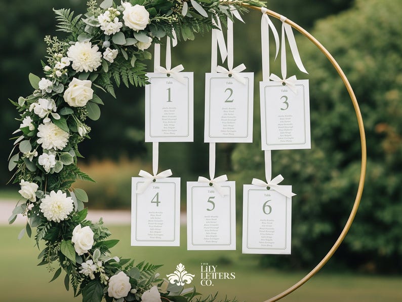 May include: A circular gold metal frame decorated with white flowers and foliage, with six white cards suspended from it. Each card is numbered and lists names. The cards are tied with white ribbons. The Lily Letters Co. logo is at the bottom.