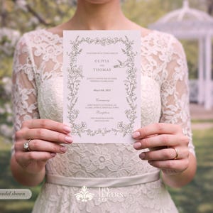 May include: A light pink wedding invitation with floral and bird illustrations. The invitation features the names "Olivia" and "Thomas" and the date "May 19th, 2024". The invitation is held by a person wearing a white lace dress.