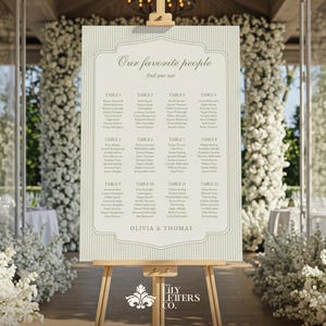 Puede incluir: Un plano de asientos de boda crema sobre un caballete de madera. El letrero tiene un borde decorativo y rayas verticales. El texto dice "Nuestra gente favorita encuentra tu asiento" y enumera nombres debajo de los números de mesa.