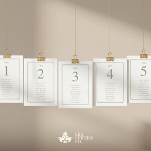 May include: Five rectangular cards with green text, each labelled with a table number from 1 to 5. The cards are suspended by gold clips and strings against a neutral background. Each card lists names.