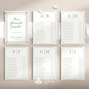 May include: A set of white wedding seating cards with green borders. The cards are displayed on a neutral surface, with a ribbon and decorative elements. The cards feature names and table assignments, with the text "Our favorite people!" on one card.