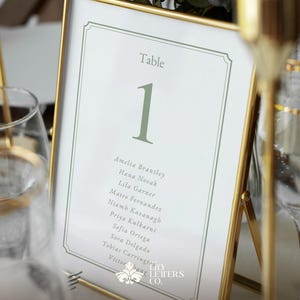 May include: A framed table number one with a gold border. The table number is in a light green colour, with a list of names below. The frame is set on a table with a glass of water and a gold candle holder.