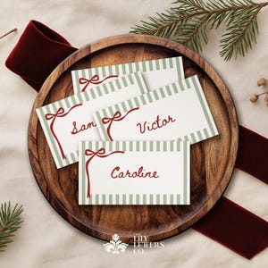 Peut inclure: Quatre cartons de noms avec des nœuds rouges et les noms "Sam", "Victor" et "Caroline", sur un plateau en bois. Les cartes ont une bordure à rayures vertes. Un ruban rouge foncé et des brins de verdure et de baies entourent le plateau.