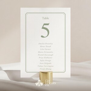 May include: A wedding table number card with the number 5 in green, and a list of names. The card is held by a gold-coloured cylindrical holder. The text "Table" is at the top.