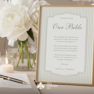 May include: A framed sign with the text "Our Bible" in elegant script, featuring a striped background and gold frame. The sign includes a message for guests to sign and highlight favorite verses. A pen and open book are in the foreground, with white flowers and candles in the background.