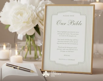 May include: A framed sign with the text "Our Bible" in elegant script, featuring a striped background and gold frame. The sign includes a message for guests to sign and highlight favorite verses. A pen and open book are in the foreground, with white flowers and candles in the background.
