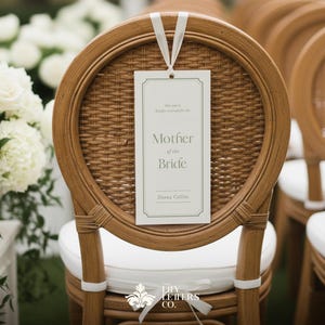 May include: A wedding ceremony chair with a woven back and white cushion. A rectangular sign reading "Mother of the Bride" hangs from the chair. White roses and hydrangeas are in the background.