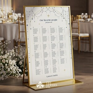 May include: A gold-framed seating chart with the text "Our favorite people find your seat." The chart lists names alphabetically. The design includes decorative stars and a floral emblem at the bottom. The chart is displayed on a gold stand.
