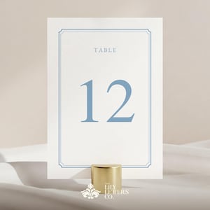 May include: A white table number card with a light blue border and the number 12 in light blue. The word "TABLE" is above the number. The card is held by a gold-colored cylindrical holder. The Lily Letters Co. logo is at the bottom.