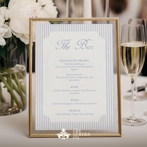 Op de afbeelding: Een goudomrand menu met de titel "The Bar", met drankjes zoals cocktails, bier, wijn en sterke drank. Het menu heeft een lichtblauwe en witte gestreepte achtergrond. Twee champagneglazen staan op de voorgrond, met een vaas met witte bloemen.