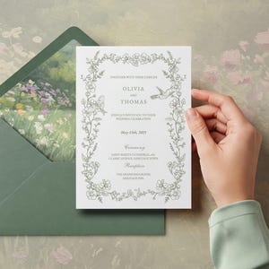 May include: A wedding invitation with floral border and hummingbird illustrations. The invitation is held by a hand and is inside a green envelope. The text on the invitation reads "Olivia and Thomas" and the date "May 15th, 2025".