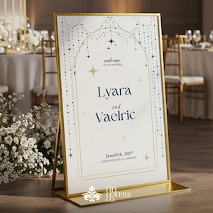 May include: A gold-framed wedding welcome sign with the names Lyara and Vaelric, and the date June 16th, 2027. The sign has a cream background with navy blue and gold celestial design elements. The sign is displayed on a gold stand.