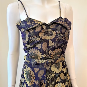 May include: A navy blue jumpsuit with a floral pattern in gold and green. The jumpsuit features spaghetti straps, a cinched waist with a matching belt, and a sweetheart neckline. The fabric has a textured appearance.