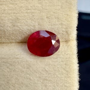 Könnte beinhalten: Ein loser, oval geformter, facettierter Edelstein in einer tiefen, leuchtenden roten Farbe. Der Stein weist auf seiner Oberfläche mehrere dreieckige und rautenförmige Facetten auf, die für ein reflektierendes, poliertes Aussehen sorgen.
