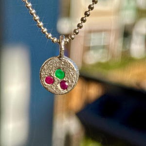 May include: A silver pendant necklace with a textured, circular pendant. The pendant features three gemstones: a green gemstone in the centre and two red gemstones below. The necklace chain is a delicate ball chain.