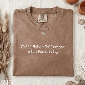 May include: Espresso-colored t-shirt with the text "Chill Vibes Collective With Positivity" in white. A silver necklace with a pendant rests on the shirt. The shirt is folded and displayed on a neutral surface.