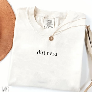 May include: Ivory-colored t-shirt with the words "dirt nerd" printed in black. A gold necklace with a small pendant rests on the shirt. The shirt has a Comfort Colors tag.