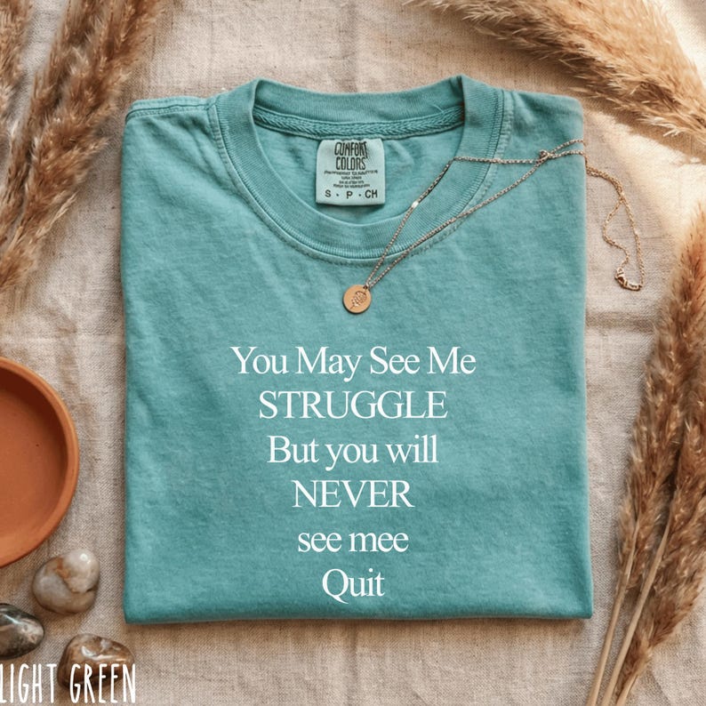 May include: Light green t-shirt with the text "You May See Me STRUGGLE But you will NEVER see mee Quit". A gold necklace with a pendant is draped over the shirt. The shirt is on a neutral surface with decorative elements.