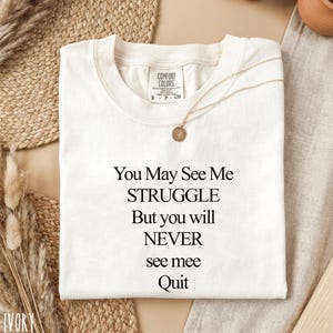 May include: Cream-coloured t-shirt featuring the quote "You May See Me STRUGGLE But you will NEVER see mee Quit" in black. A gold necklace with a circular pendant is on the shirt. The shirt is a Comfort Colors brand, size S.