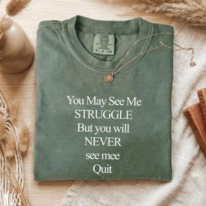 May include: An olive green t-shirt with the text "You May See Me STRUGGLE But you will NEVER see mee Quit" in white. A gold necklace with a pendant rests on the shirt. The shirt is folded on a light-coloured surface.