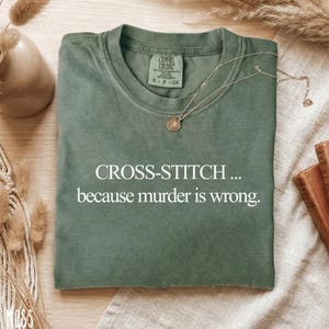 May include: A sage green t-shirt with the text "CROSS-STITCH... because murder is wrong." A gold necklace with a pendant rests on the shirt. The shirt has a Comfort Colors tag.