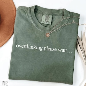 May include: A moss green t-shirt with the white text "overthinking please wait...". The shirt has a crew neck and short sleeves. A silver necklace with a small pendant is draped across the shirt.