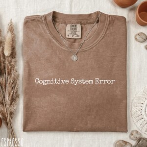 Może przedstawiać: Koszulka w kolorze espresso z białym napisem "Cognitive System Error". Koszulka ma okrągły dekolt i krótkie rękawy. Srebrny naszyjnik z wisiorkiem spoczywa na koszulce. Koszulka jest z Comfort Colors.
