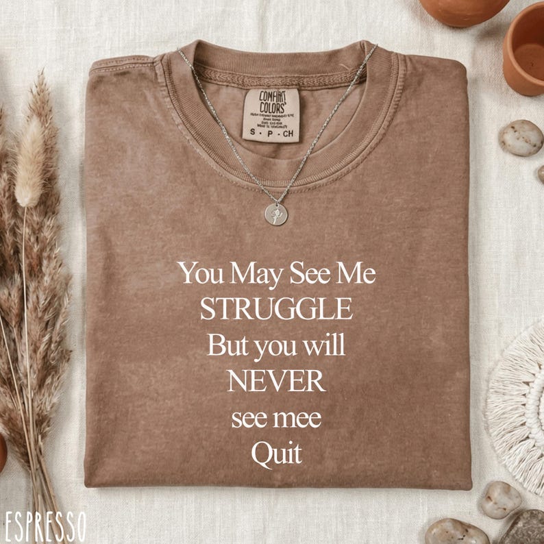 May include: Espresso-coloured t-shirt with the text "You May See Me STRUGGLE But you will NEVER see mee Quit." The shirt has a crew neck and short sleeves. A silver necklace with a pendant is on the shirt.