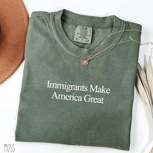 Puede incluir: Camiseta verde oliva con el texto blanco "Immigrants Make America Great". La camiseta tiene cuello redondo y mangas cortas. Un collar plateado con un pequeño colgante descansa sobre la camiseta. Un sombrero marrón es visible en el fondo.