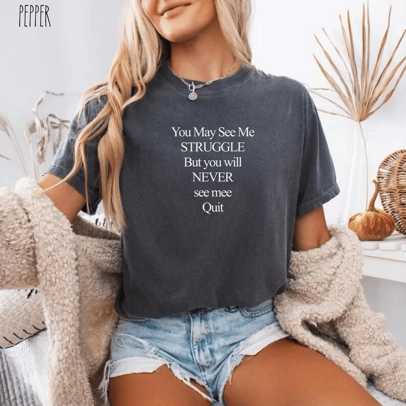 May include: Dark grey t-shirt with the text "You May See Me Struggle But you will NEVER see mee Quit" in white. The shirt is a short-sleeved crew neck style.