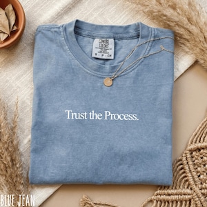 May include: A blue jean colored t-shirt with the phrase "Trust the Process" printed in white. The shirt is folded and has a gold necklace with a circular pendant. The shirt is from Comfort Colors.