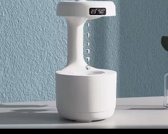 May include: White humidifier with a digital display showing 23° 50%. The humidifier has a round top and a base with a circular opening. The humidifier is emitting a fine mist.