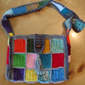 May include: A handmade crochet shoulder bag with a patchwork design. The bag features a variety of colorful squares in shades of red, orange, green, blue, and pink, framed by a gray border. It has a matching strap and a gray tassel.