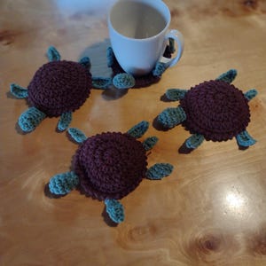 Może przedstawiać: Szydełkowe podstawki w kształcie żółwi w odcieniach bordowego i turkusowego. Podstawki mają wzór żółwia, z bordową skorupą i turkusowymi płetwami. Biały kubek spoczywa na jednej z podkładek, a trzy inne leżą na drewnianej powierzchni.