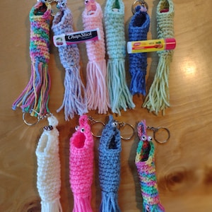 May include: A collection of 11 crocheted lip balm holders with keychains. The holders are in various colours, including pink, blue, green, white, and multi-coloured. Each holder has a small crocheted tail and a pair of googly eyes.