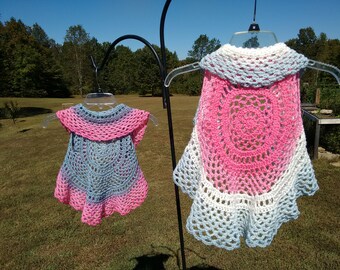 Op de afbeelding: Twee gebreide vesten, een in roze en blauw en de andere in wit en blauw, hangen aan hangers tegen een groene grasachtige achtergrond.