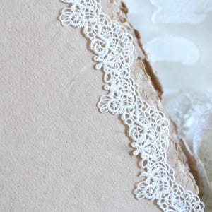 May include: A thick, light beige paper book with a rough, deckled edge. A strip of intricate white floral lace trim is attached along the vertical edge of the page. A pair of small metal scissors rests near a pile of white lace fabric.