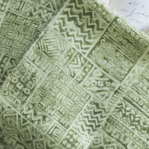 May include: Green fabric with a white geometric pattern. The pattern is made up of squares and rectangles with different designs. The fabric has a textured surface.