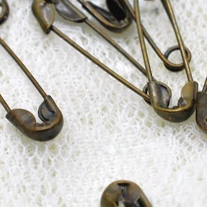 May include: A collection of antique bronze-toned metal safety pins. Each pin features a coiled spring mechanism at one end and a protective clasp at the other. The pins are arranged in a scattered formation, showcasing their metallic finish.