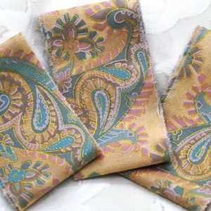 May include: Three strips of fabric featuring a gold background with a repeating paisley pattern in shades of teal, pink, and light blue. The edges of the fabric strips are frayed, and the design includes thin gold metallic outlines throughout.